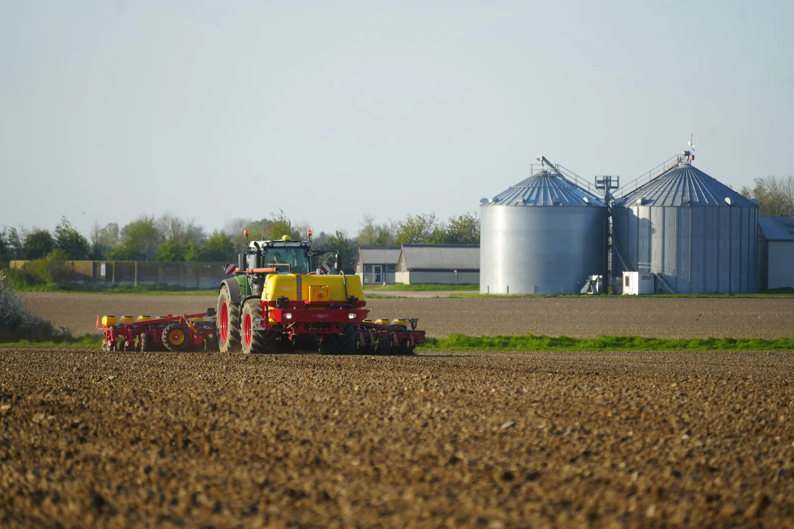 Fendt tractor drilling with a Väderstad planter beside grain silos